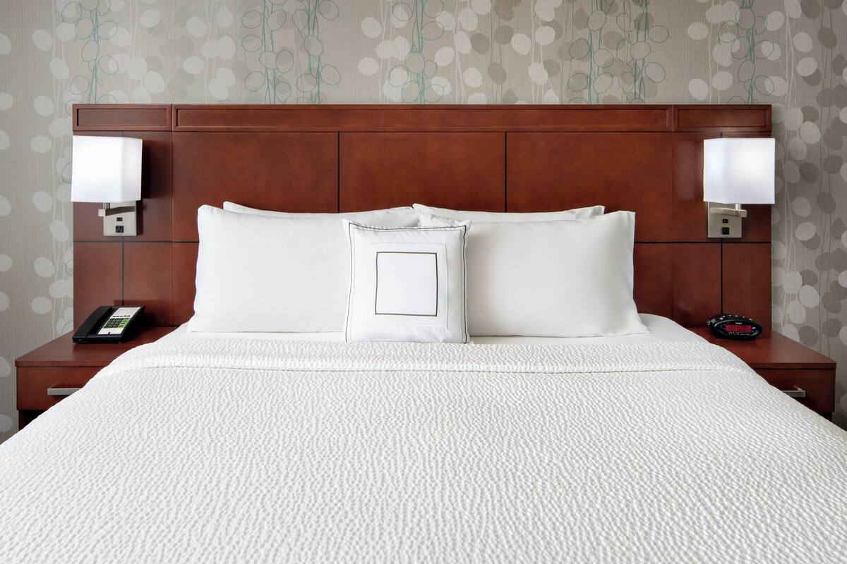 A neatly made bed with white bedding, two bedside lamps, a phone, and a clock on a wooden nightstand.