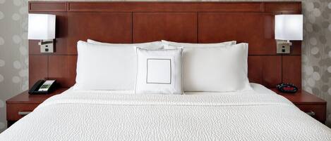Una cama bien hecha con ropa blanca, dos lámparas de noche, un teléfono y un reloj sobre una mesita de noche de madera.