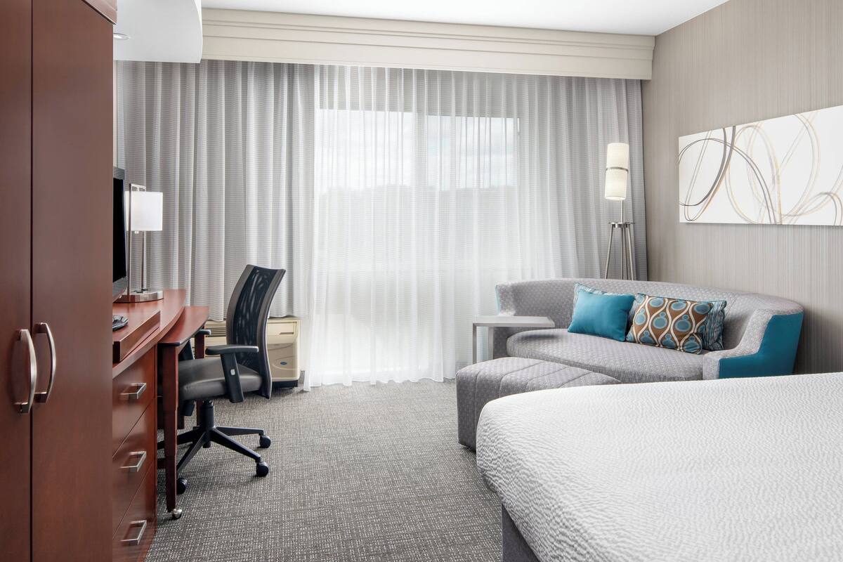 A hotel room with a bed, a desk, a chair, a lamp, and a large window with curtains.