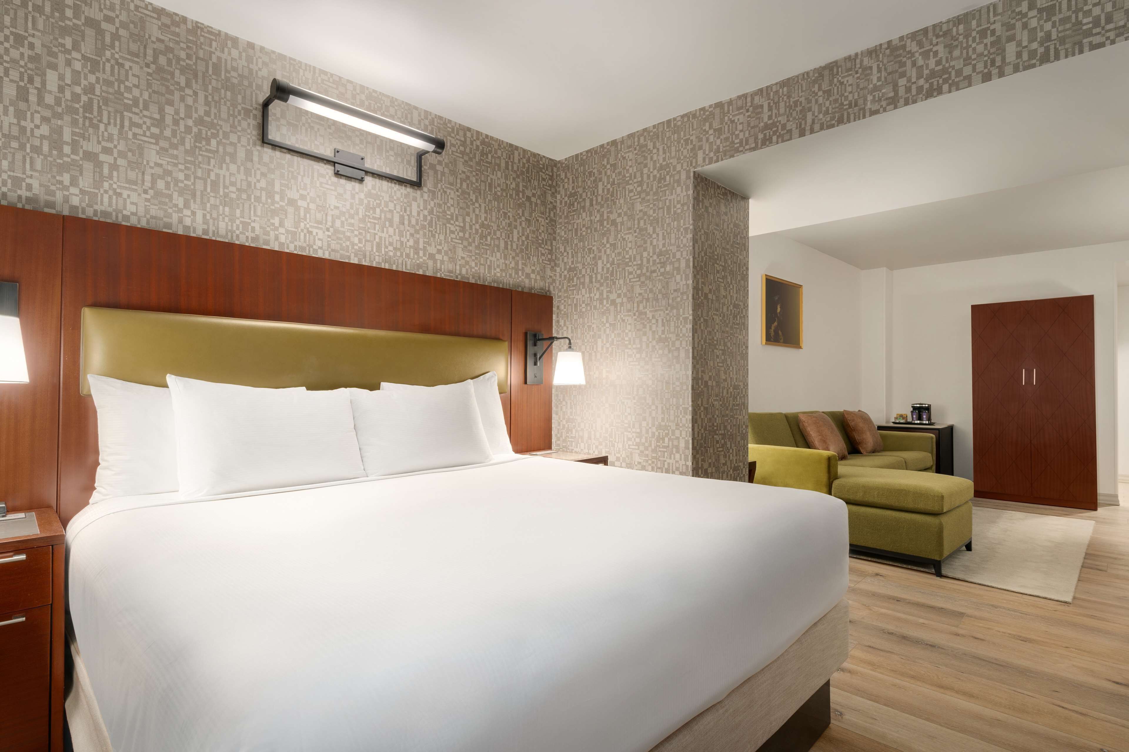 A hotel room with a large bed, a wooden headboard, a nightstand, a lamp, a green sofa, and a wardrobe.