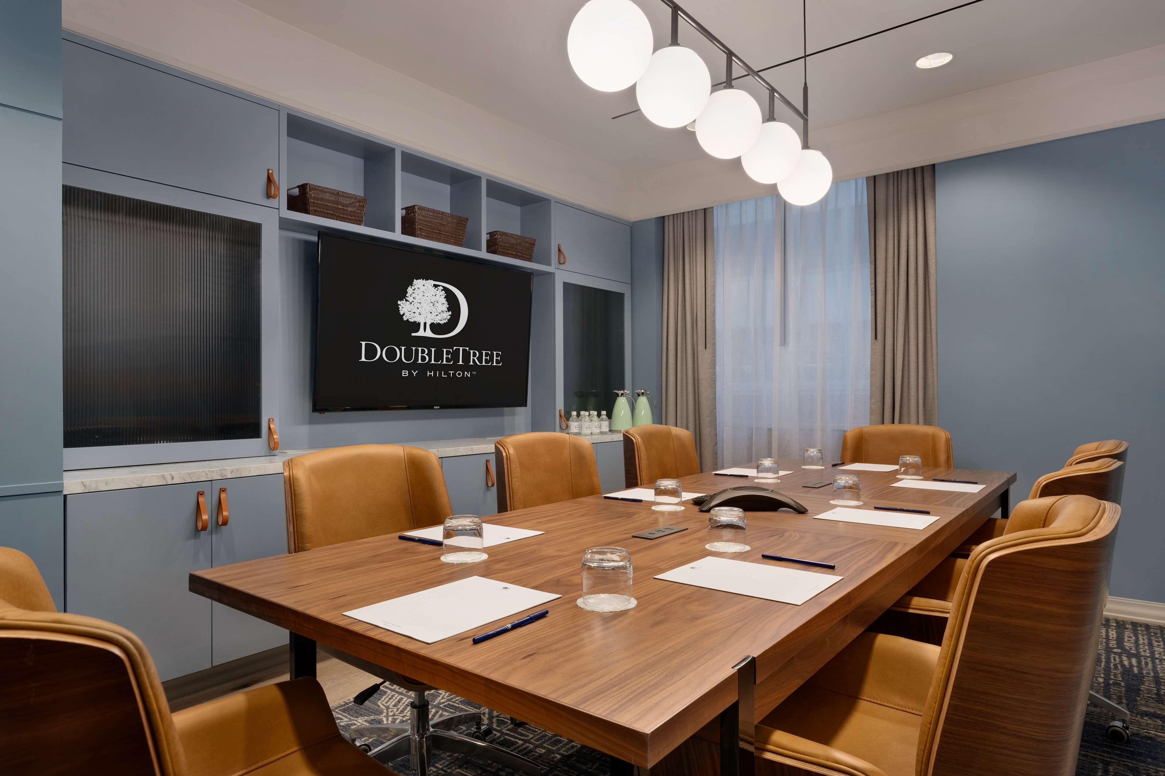 A conference room with a long table, brown chairs, a large screen displaying 'DOUBLETREE by Hilton', and a modern chandelier.