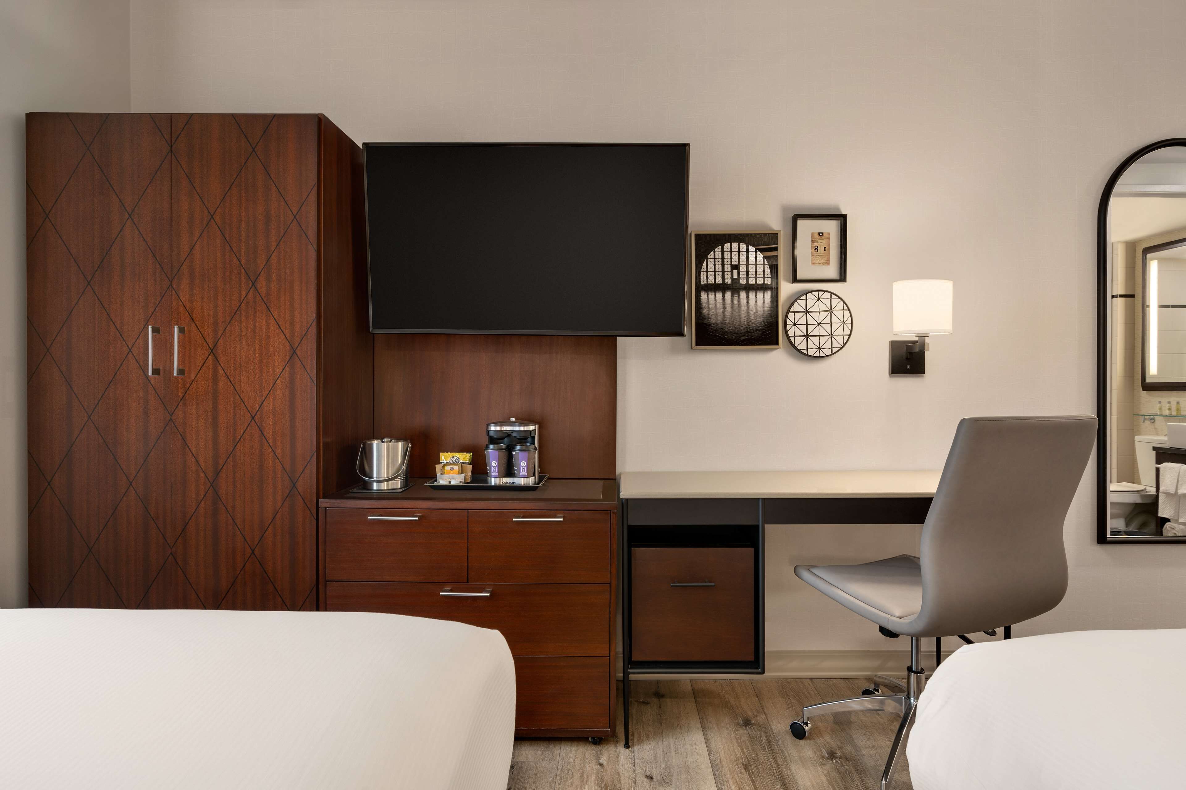 Habitación de hotel con cama, escritorio y silla, armario, televisión de pantalla plana, cafetera y lámpara de pared.