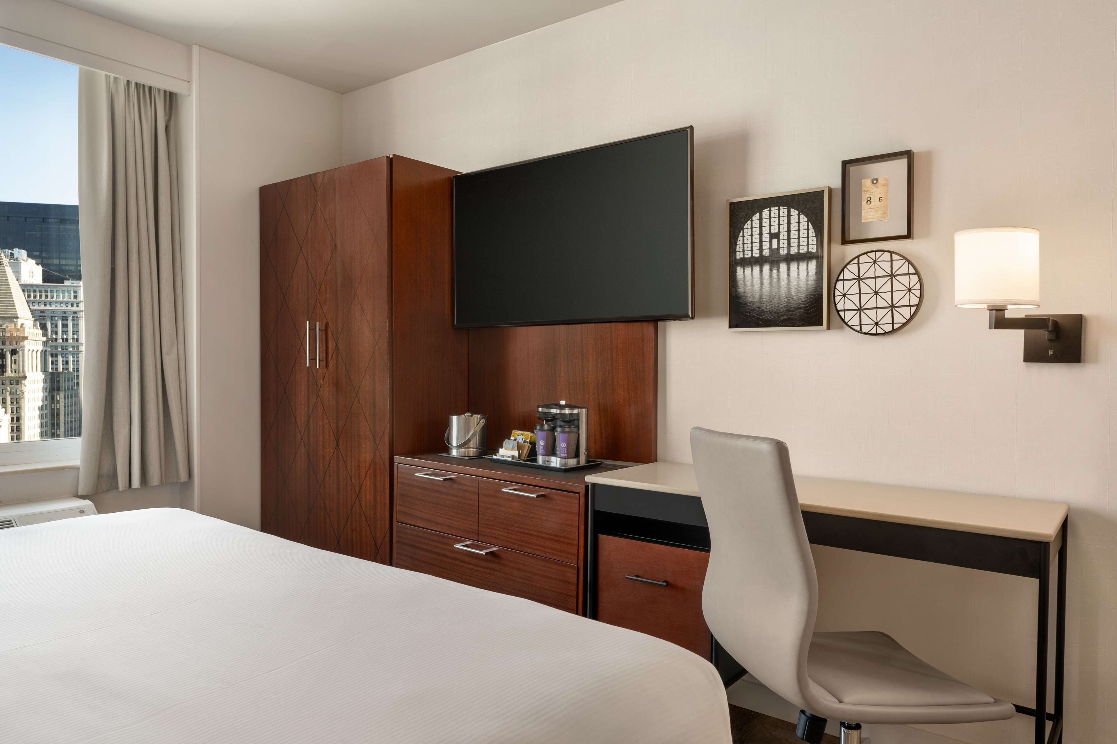 A hotel room with a bed, a desk with a chair, a wardrobe, a flat-screen TV, and artwork on the wall.