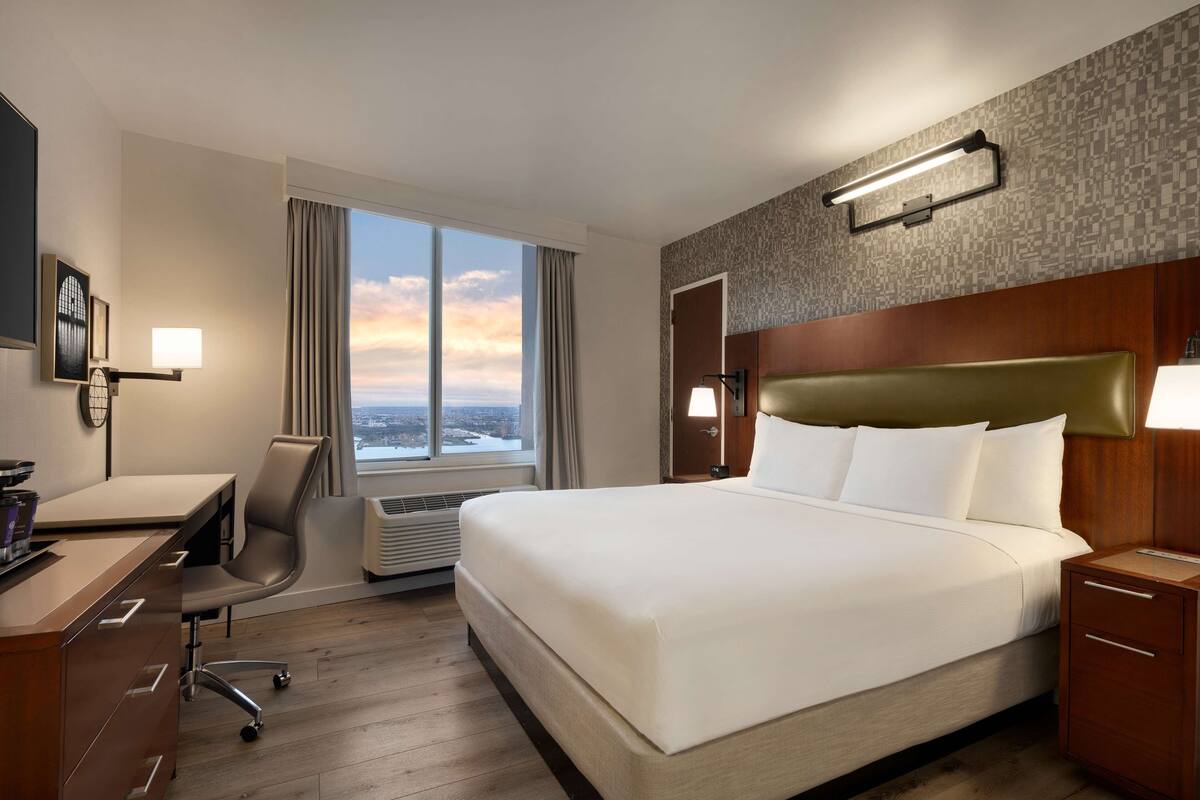 Hotellihuone, jossa on suuri sänky, työpöytä, tuoli ja ikkunasta avautuva näkymä kaupungin ylle.