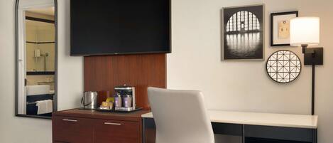 A hotel room with a bed, a desk with a chair, a television, a coffee maker, a mirror, and artwork on the wall.