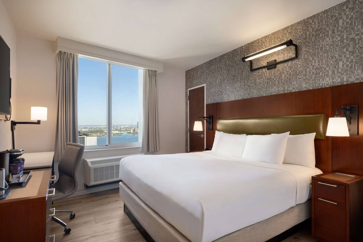 Hotellihuone, jossa on suuri sänky, työpöytä tuolilla, televisio ja ikkuna, jossa on verhot.
