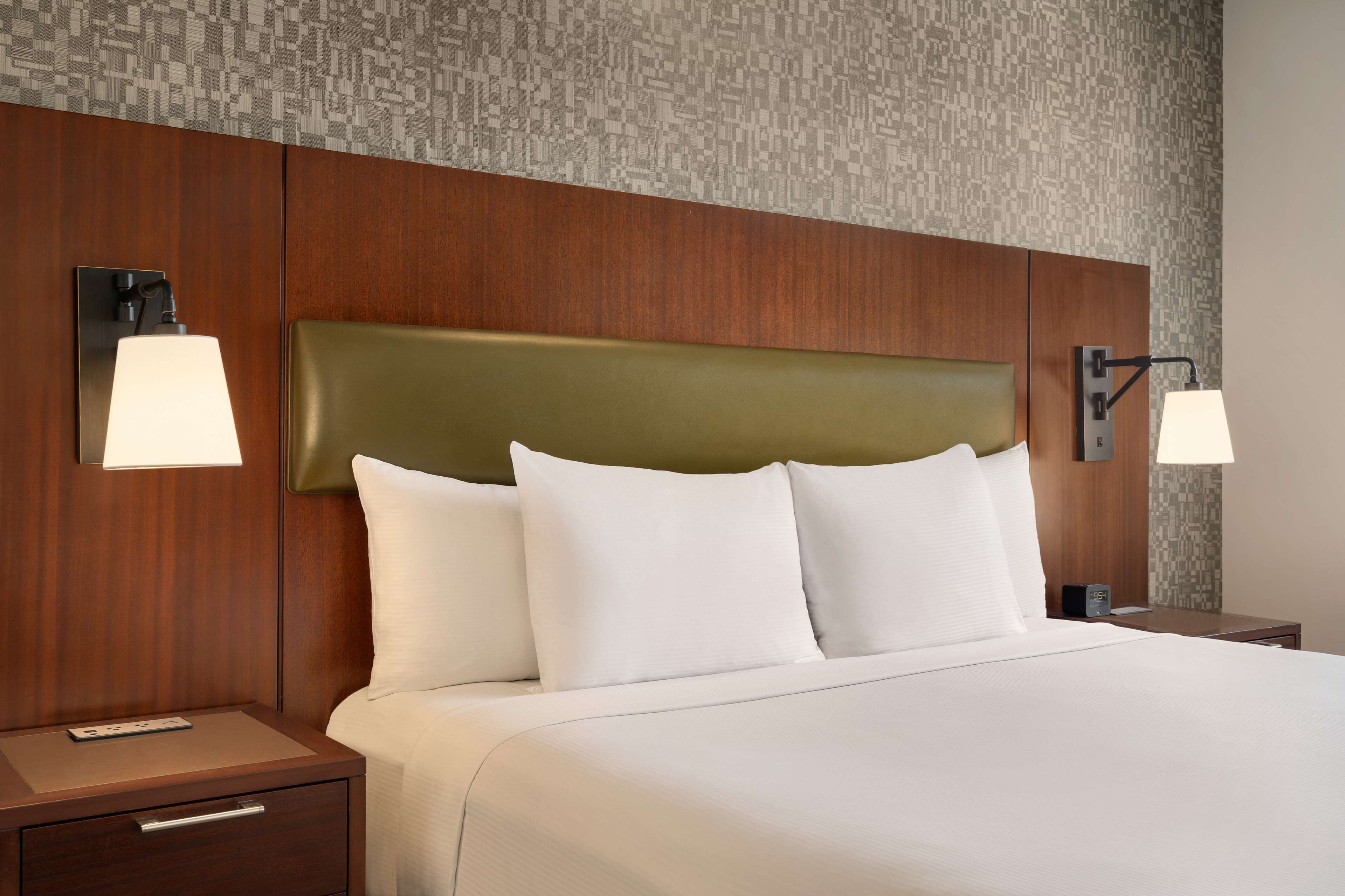 A hotel room with a large bed, wooden headboard, two bedside tables with lamps, and a small drawer.
