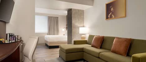 A hotel room with a sofa, a desk, a chair, a bed, a TV, a lamp, and a painting on the wall.