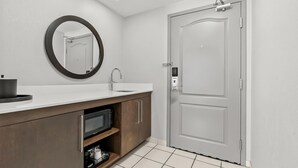 A modern bathroom with a white vanity, a round mirror, a built-in microwave, and a door with a handle.