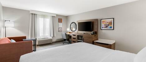 Habitación de hotel con una cama grande, un escritorio, una silla, un televisor, un espejo y una ventana con cortinas.