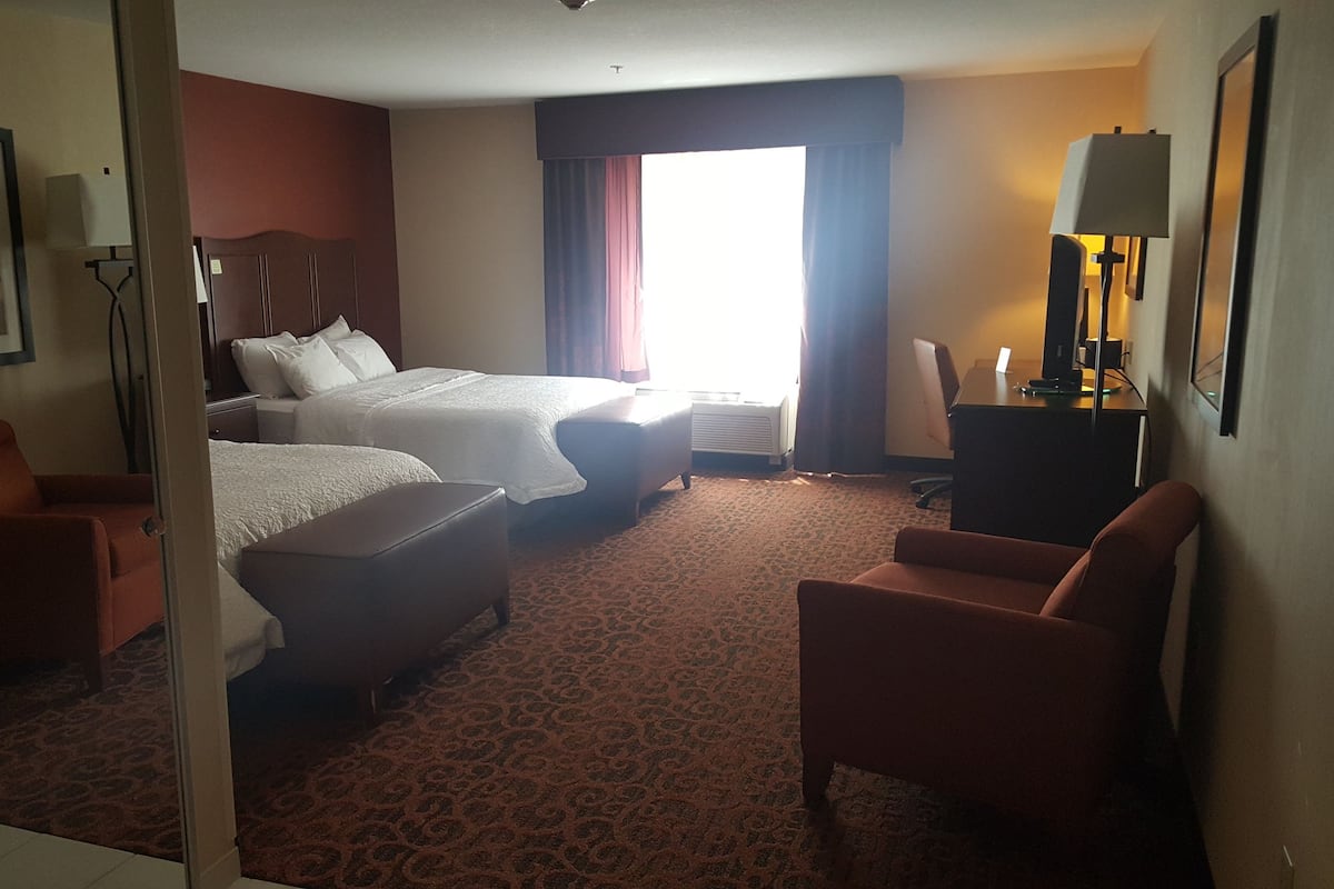 A hotel room with two beds, a desk, a chair, and a large window with curtains.