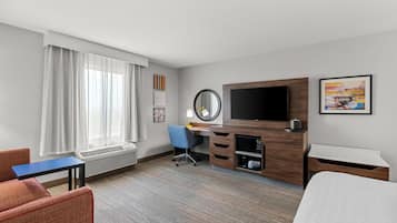 A hotel room with a bed, a desk with a chair, a TV, a window with curtains, and artwork on the walls.
