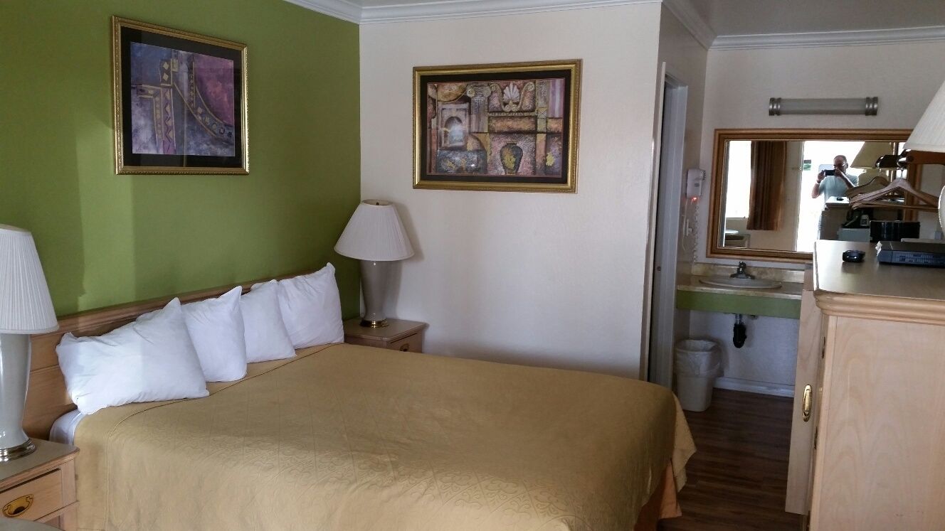 Habitación de hotel con una cama grande, dos cuadros en las paredes, una mesita de noche con lámpara, un pequeño atril, un espejo, un aparador y un baño visible a través de una puerta abierta.
