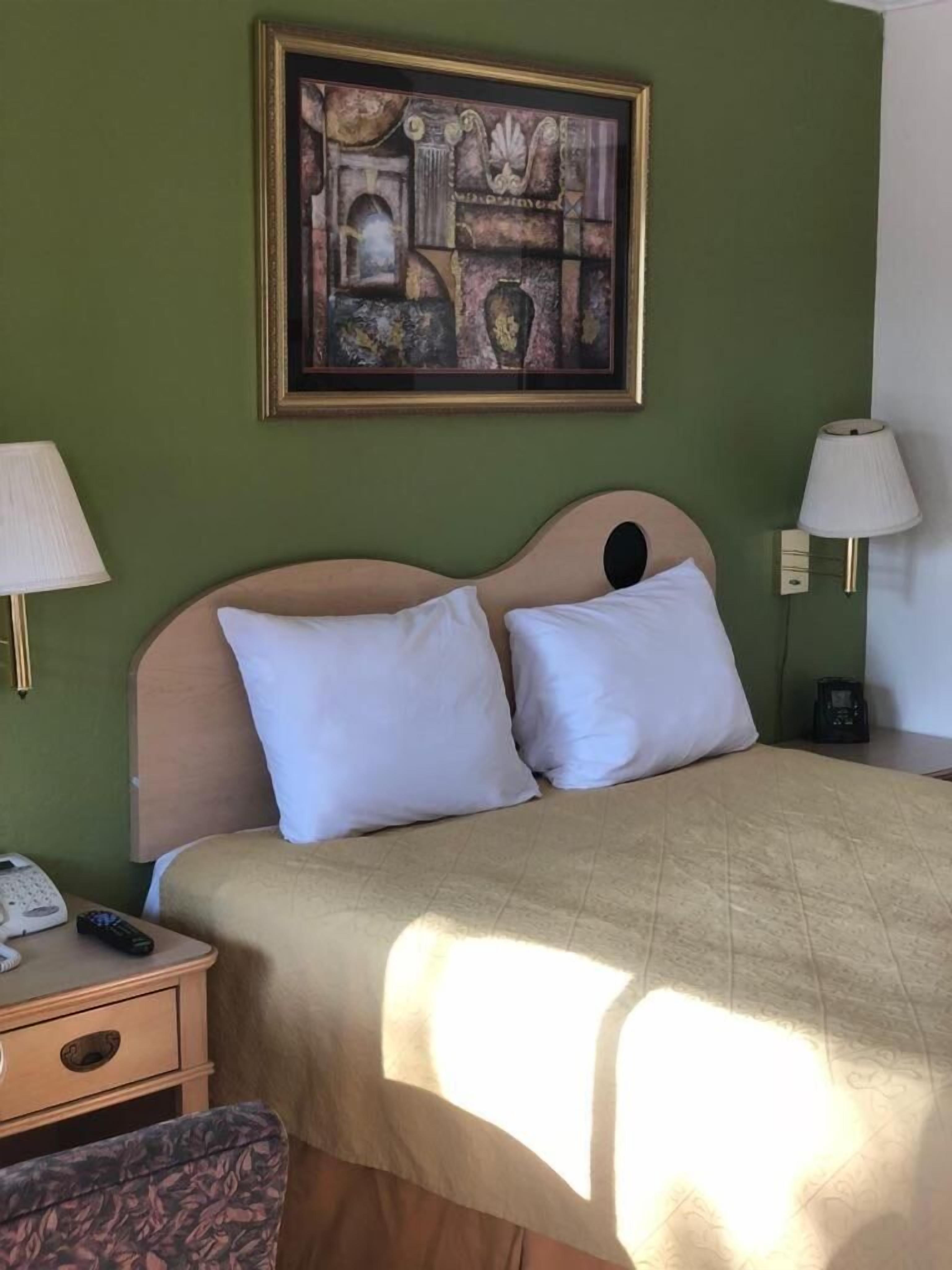 Une chambre d’hôtel comprenant un lit, deux lampes de chevet, une table de chevet avec un téléphone et une télécommande, une chaise et une œuvre d’art encadrée accrochée au mur.