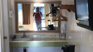 Quarto compacto com piso de madeira, uma cama, escrivaninha, espelho, televisão e uma pequena cozinha.