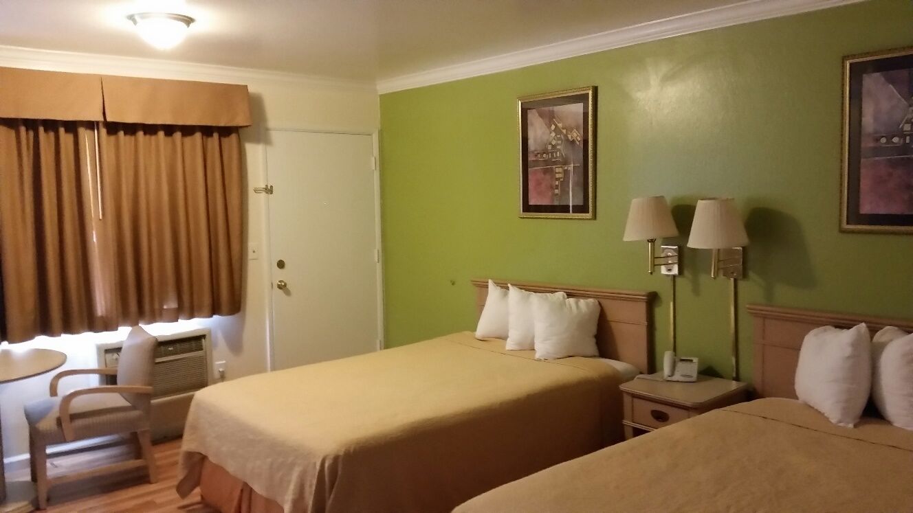A hotel room with two beds, a desk, and a chair. There are two wall-mounted lamps and a painting on the wall.