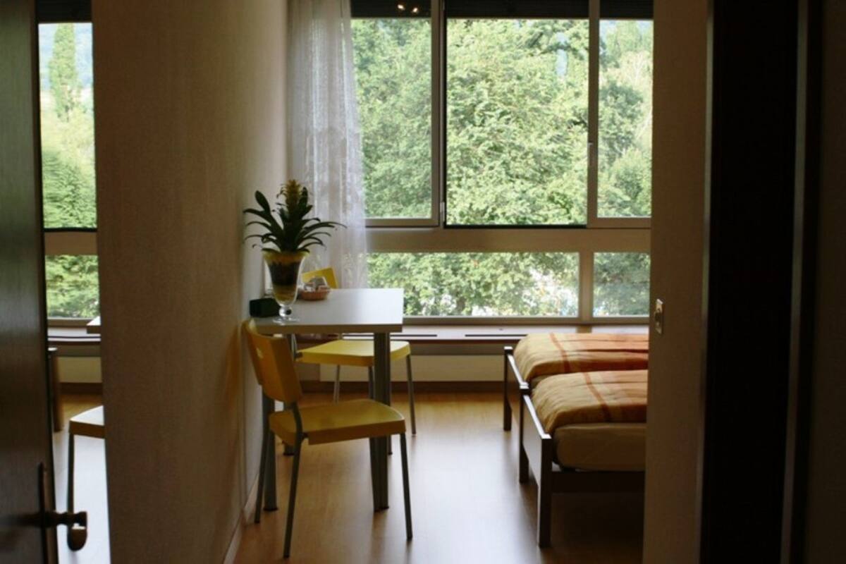 A room with a bed, a table with a chair, and a window with a view of greenery.