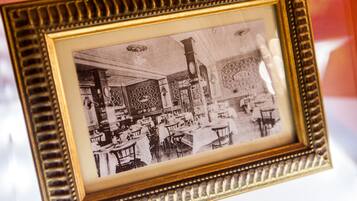 Fotografia a preto e branco de uma sala de jantar com mesas, cadeiras, um relógio e padrões nas paredes.