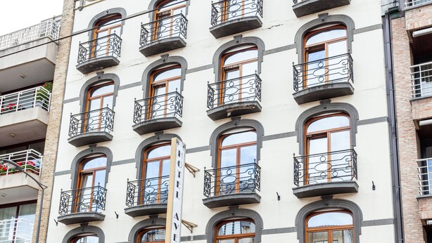 Un immeuble à plusieurs étages, doté d’une façade blanche, de nombreux balcons ornés de rampes en fer forgé, et d’une entrée au niveau du sol avec un banc en bois et des plantes en pot.