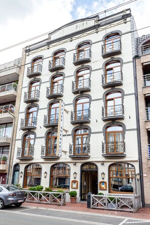 A multi-story building with a white facade, multiple balconies with wrought iron railings, and a ground-level entrance with a wooden bench and potted plants. - Avenue Hotel by F-Hotels (Blankenberge)
