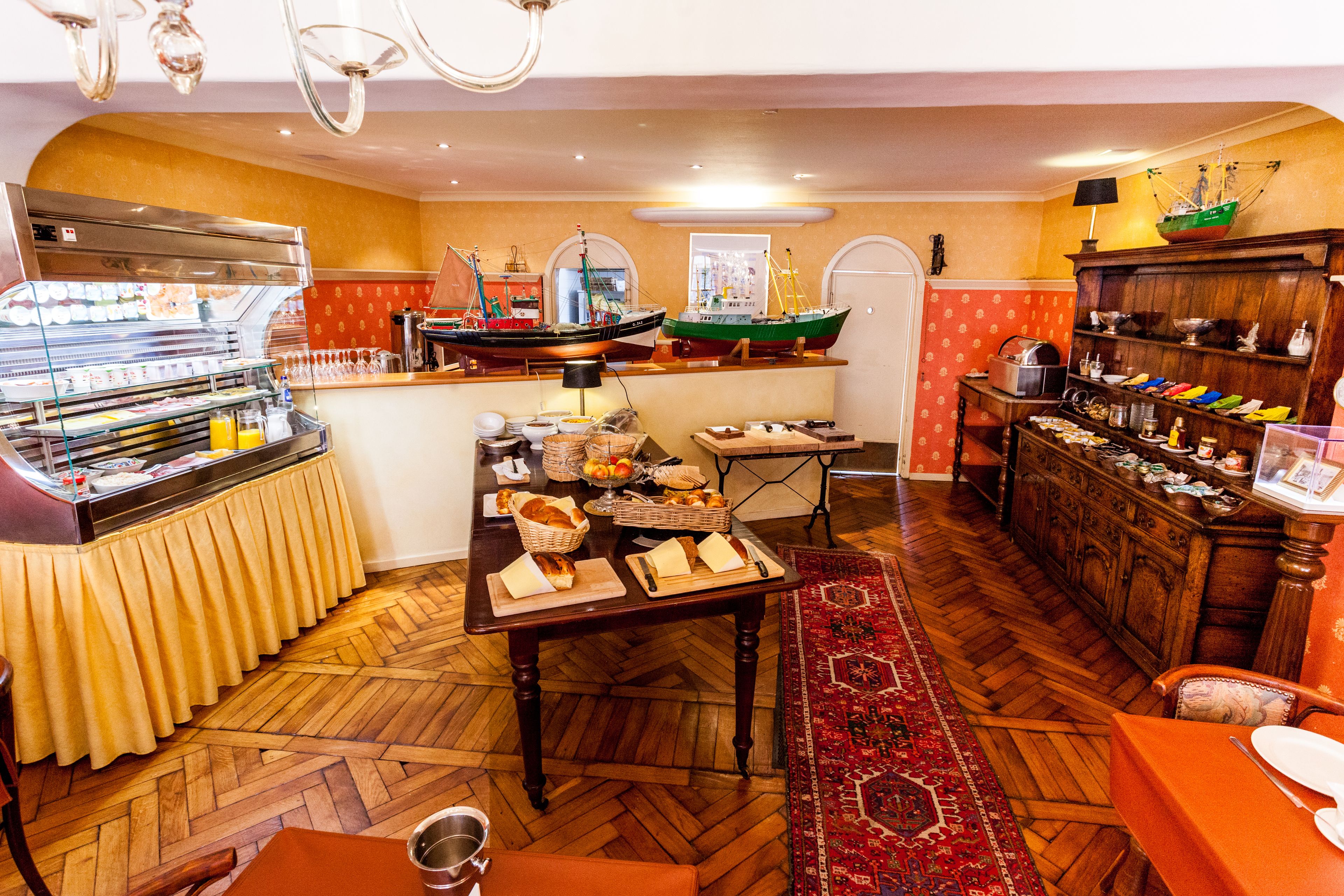 A restaurant with a wooden herringbone floor, a buffet with various food items, and a display of model ships.