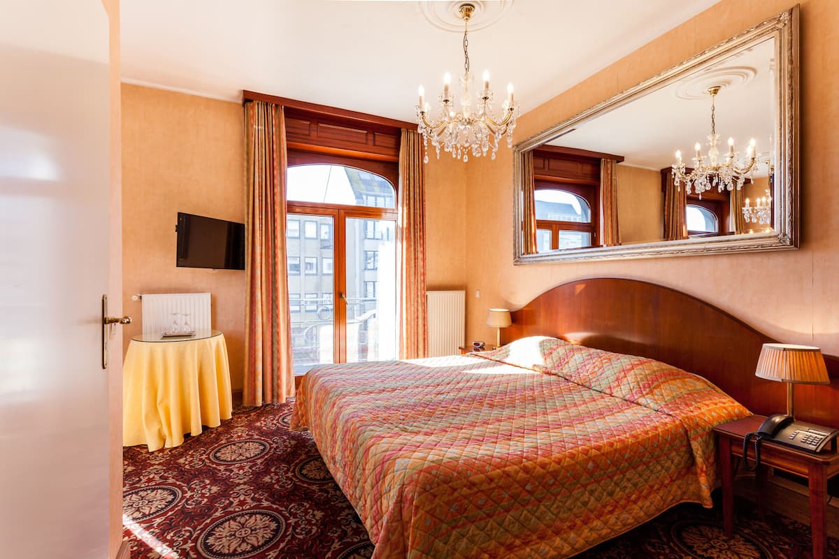 Ein Hotelzimmer mit einem großen Bett, einer Kandelaber, einem Spiegel, einem Nachttisch mit Lampe und Blick auf die Stadt durch das Fenster.