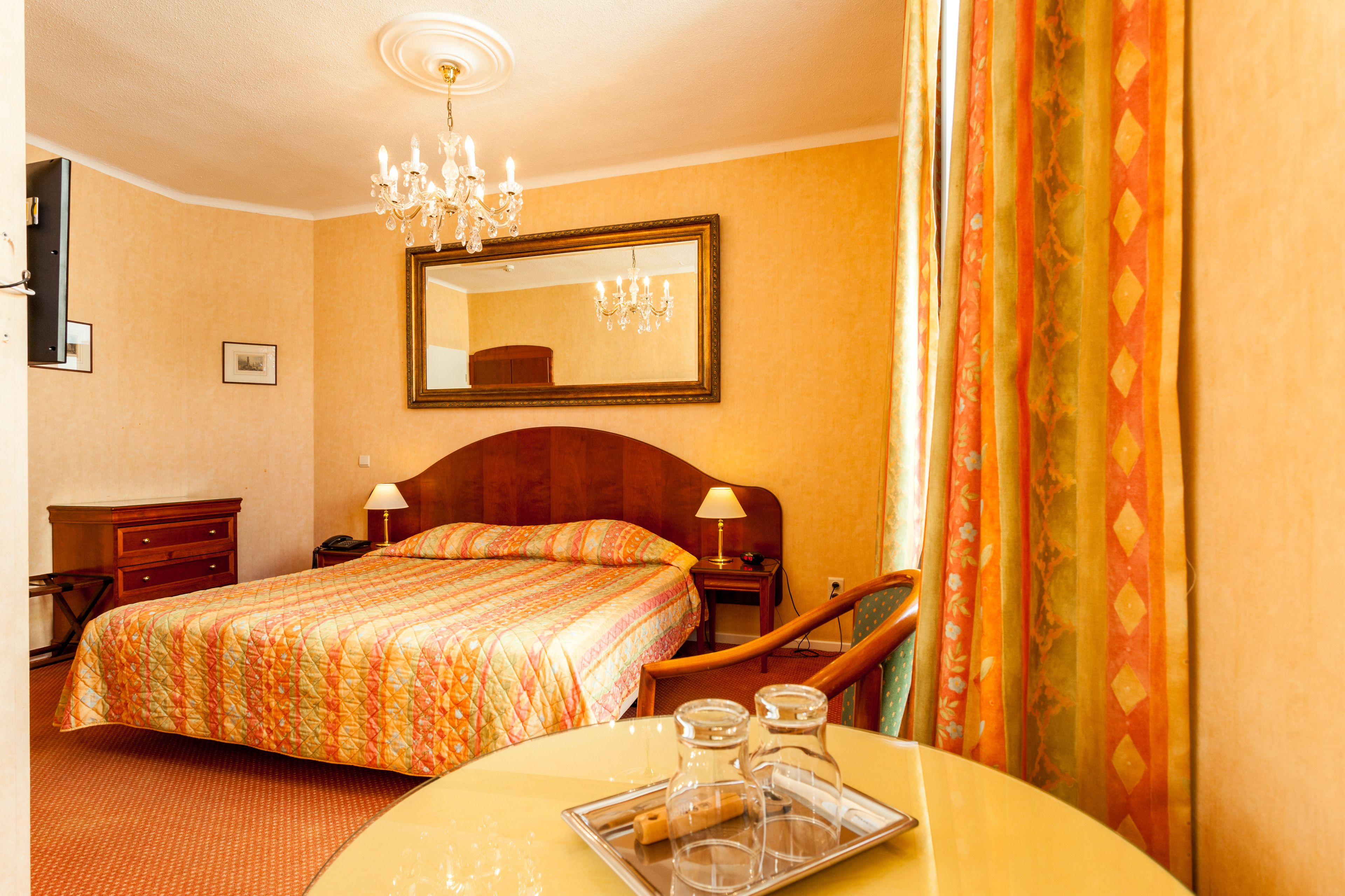 A hotel room with a large bed, a wooden headboard, a chandelier, a mirror, a nightstand, a lamp, and a tray with glasses and a bottle.