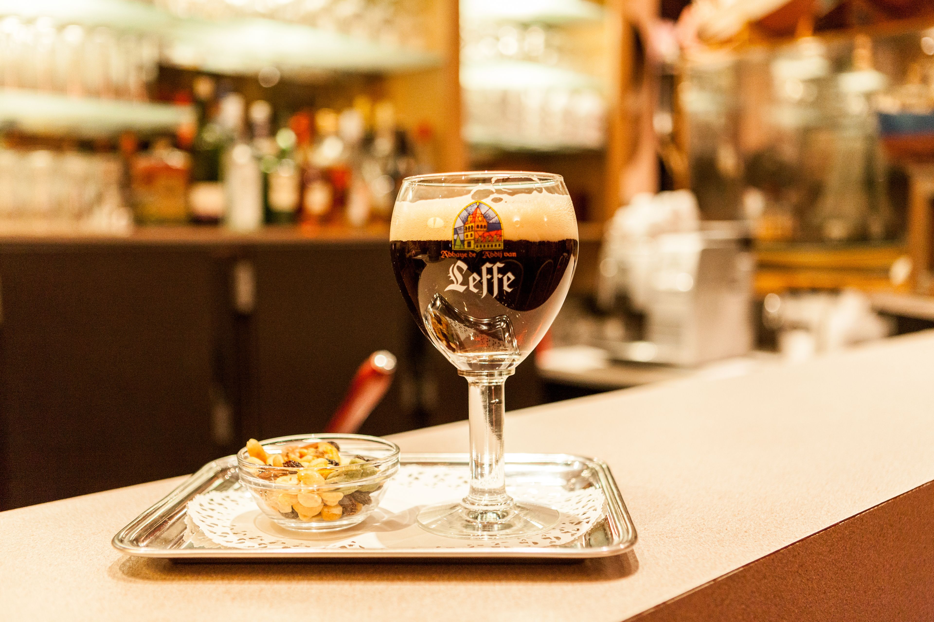 A glass of dark beer with a logo on the side, placed on a tray with a small bowl of nuts beside it.