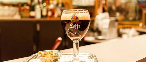 A glass of dark beer with a logo on the side, placed on a tray with a small bowl of nuts beside it.
