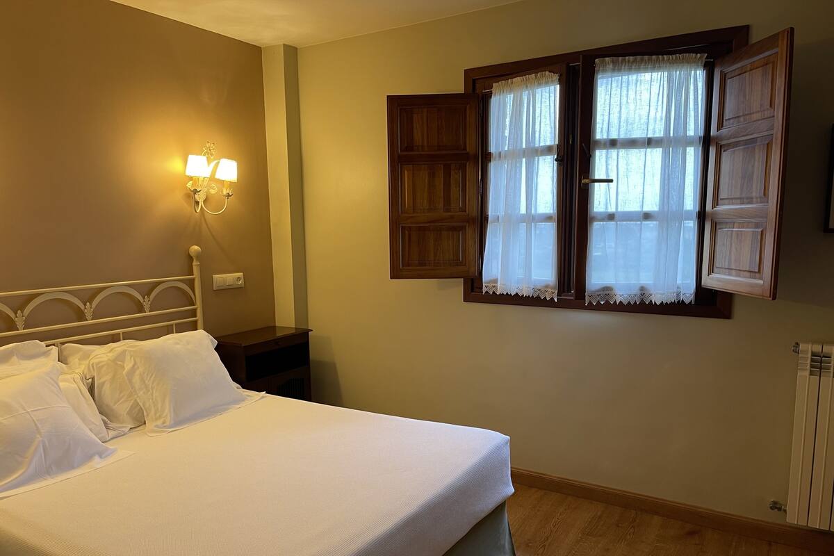 A neatly made bed with white linens, a bedside table with a lamp, and a window with wooden shutters.