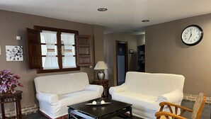 A living room with two white sofas, a wooden coffee table, a wicker chair, a wall clock, and a window with curtains. - Casona de Cefontes (Gijon)