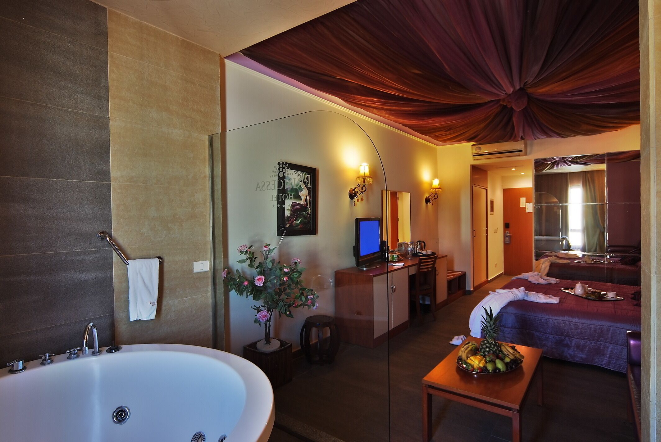 Een hotelkamer met een groot bad, een bureau met een computer, een bed en een klein tafeltje met fruit.