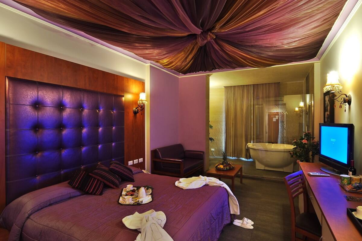 A hotel room with a tufted headboard, a bed with pillows, a bedside table with a lamp, a desk with a computer, and a bathtub in the background.