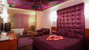 A bedroom with a tufted headboard, a bed with a rose petal arrangement, a vanity with a mirror, and a bathroom with a bathtub and shower.