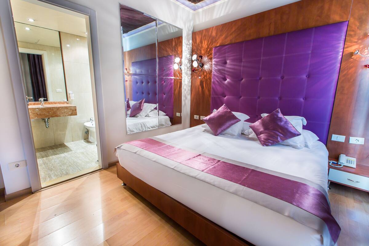 A modern hotel room with a large bed, a glass shower enclosure, and a wooden vanity.