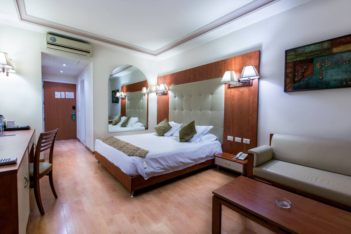 A hotel room with a bed, a desk, a chair, a sofa, and a painting on the wall.