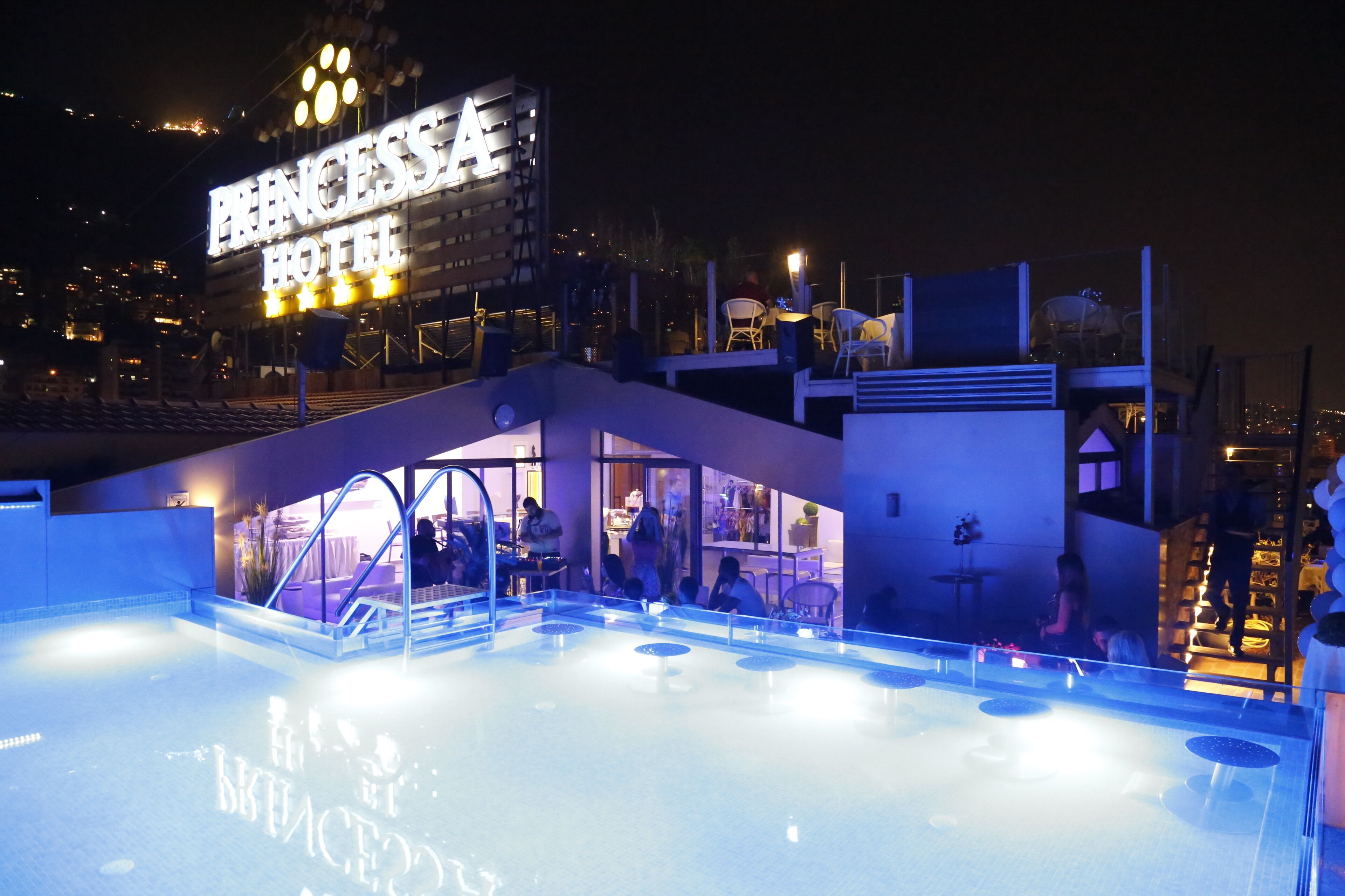 A nighttime view of a hotel with a pool area, outdoor seating, and a prominent illuminated sign.