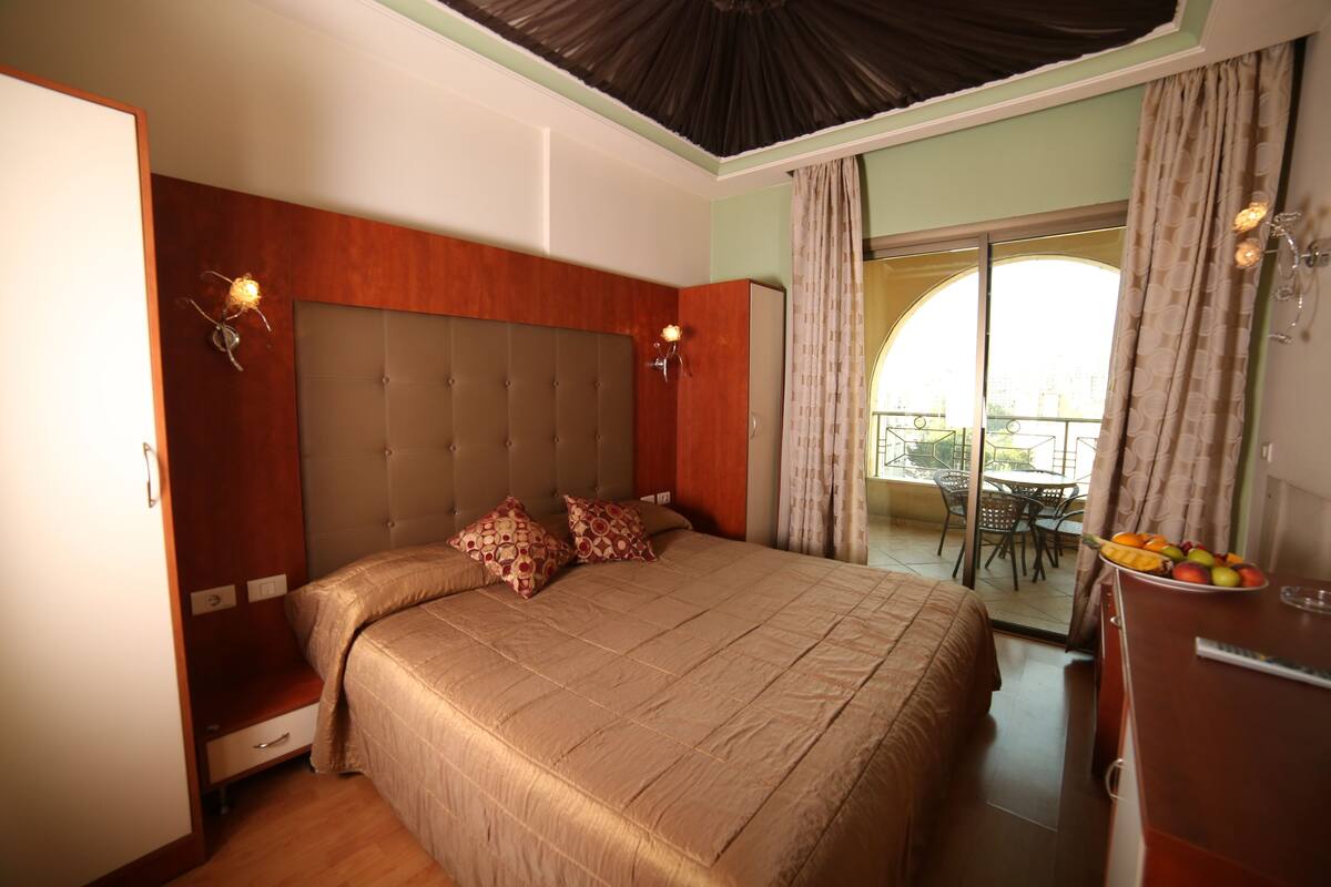 A bedroom with a large bed, a wardrobe, a desk with fruit, and a balcony with a table and chairs.