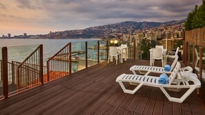 Une terrasse en bois, dotée de chaises longues blanches et d’une rambarde offrant une vue sur une marina et le paysage urbain.