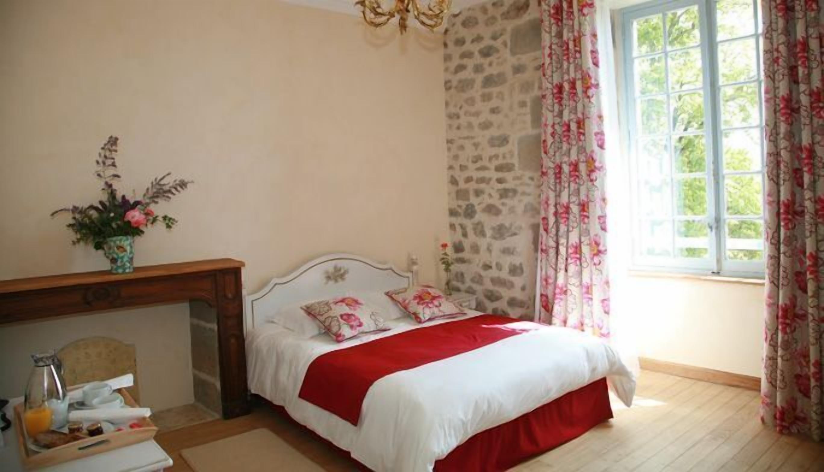 A bedroom with a bed, a wooden dresser, a vase with flowers, a window with curtains, and a stone wall.
