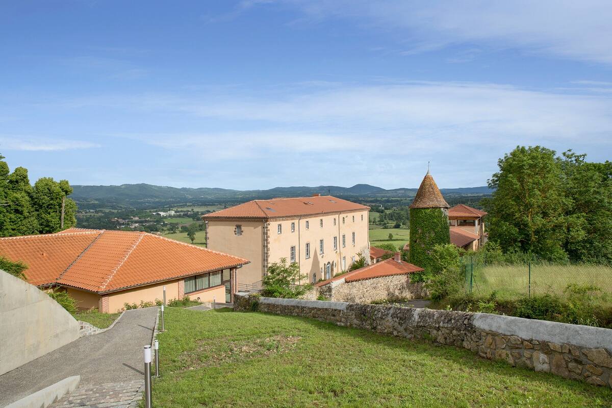 A building with a red-tiled roof and a stone tower, surrounded by greenery and hills.