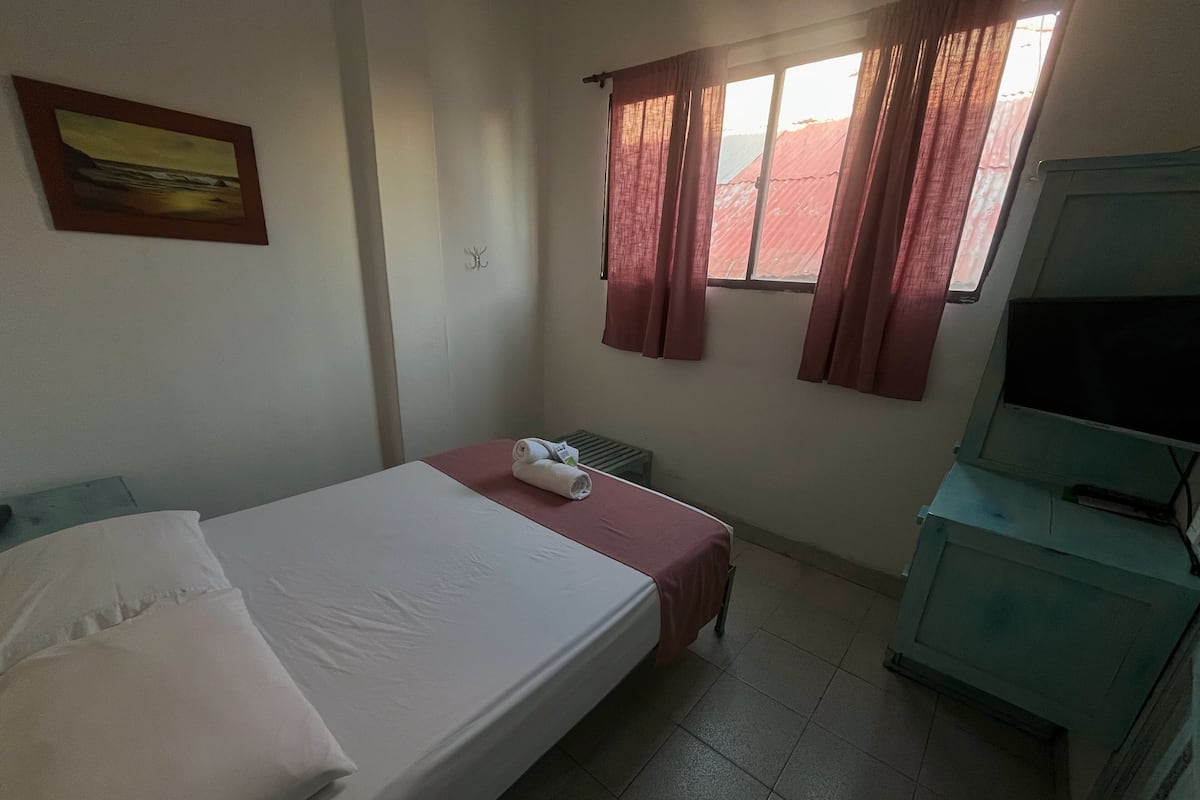 A hotel room with a bed, a desk, a chair, a radiator, and a window with curtains.