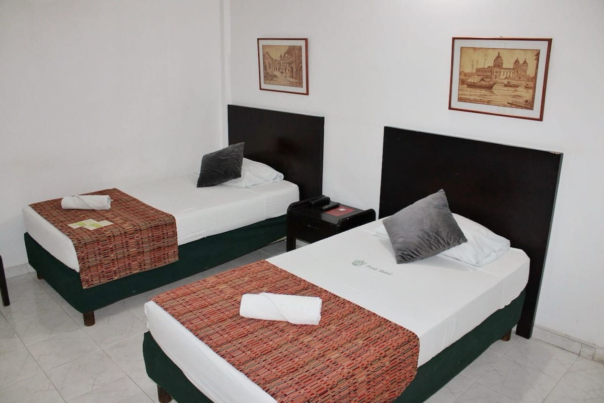 Two single beds with dark headboards, white bedding, and a red patterned bedspread. Each bed has a pillow and a folded towel. There are framed pictures on the wall above the beds.