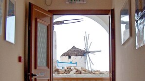 An open door leading to a balcony with a view of a traditional building and a windmill. - The Windmills (Ios)