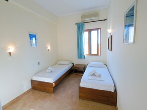 Une petite chambre propre, avec deux lits simples, une fenêtre avec des rideaux, un climatiseur fixé au mur et une petite table.