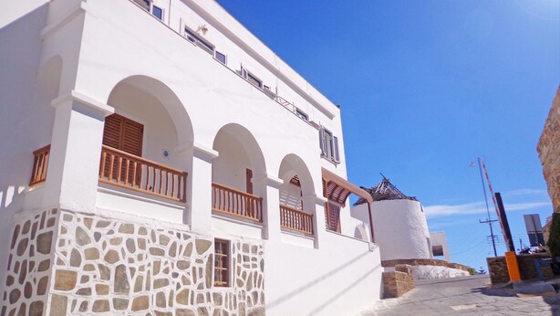A white building with stone facade and arched windows, a paved street, and a clear blue sky. - The Windmills (Ios)