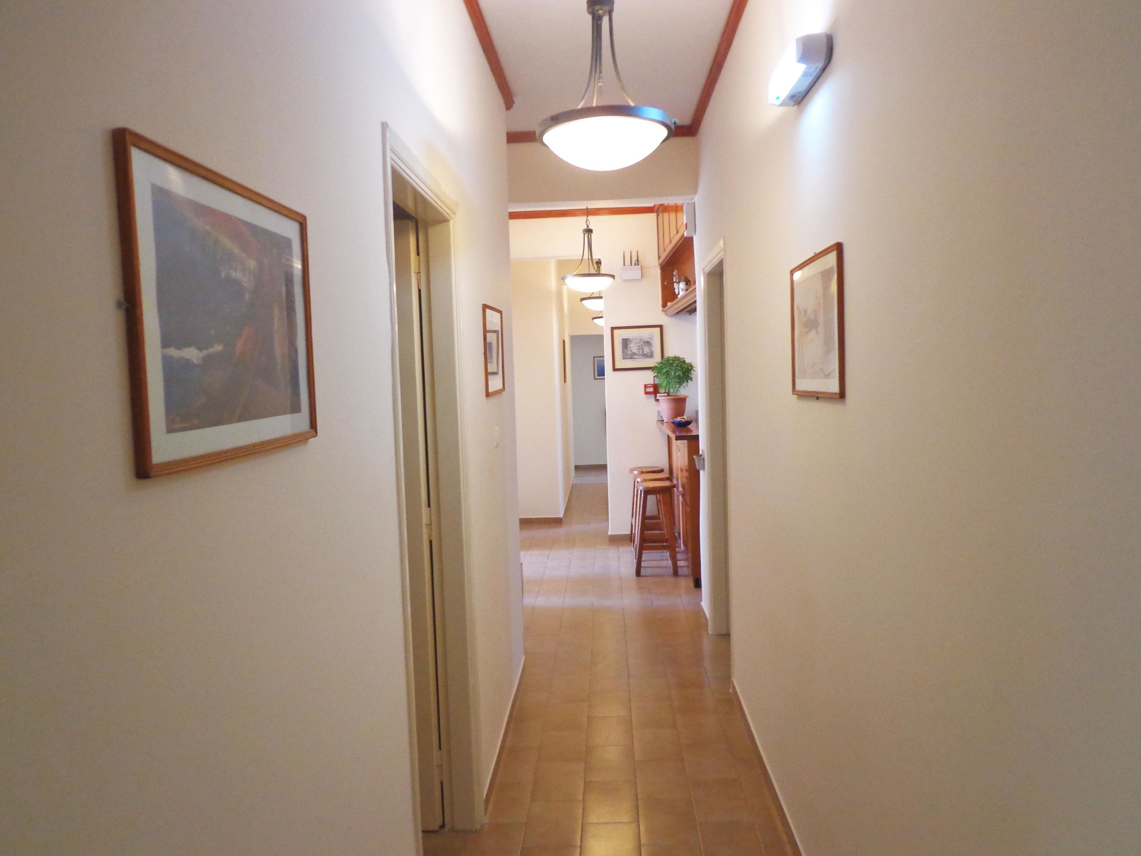 A narrow hallway with framed artwork on the walls, a wooden door, and a kitchen area with bar stools and a potted plant.