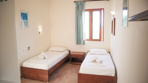 A room with two wooden beds, a small bedside table, a wall-mounted lamp, a window with a curtain, and a framed picture on the wall. - The Windmills (Ios)