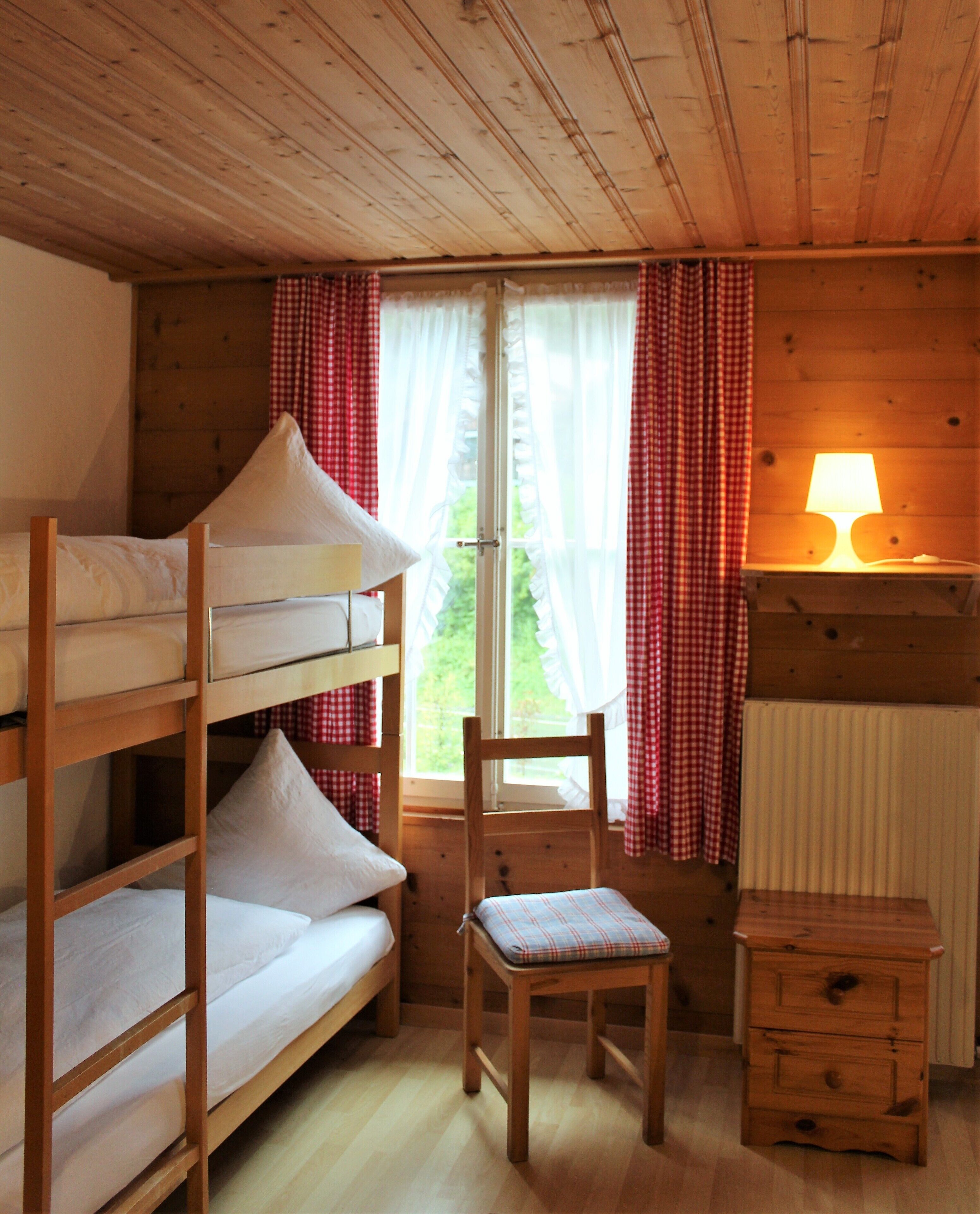 A wooden cabin room with bunk beds, a chair, a small table, and a window with curtains.