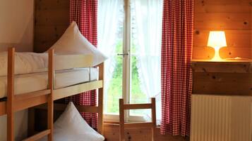 A wooden cabin room with bunk beds, a chair, a small table, and a window with curtains.